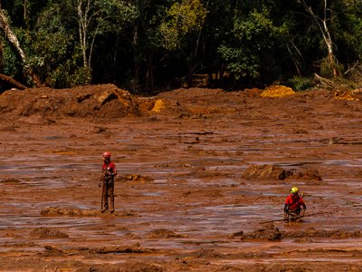 Two people in red shirts and helmets stand in red mud, there are trees in the background